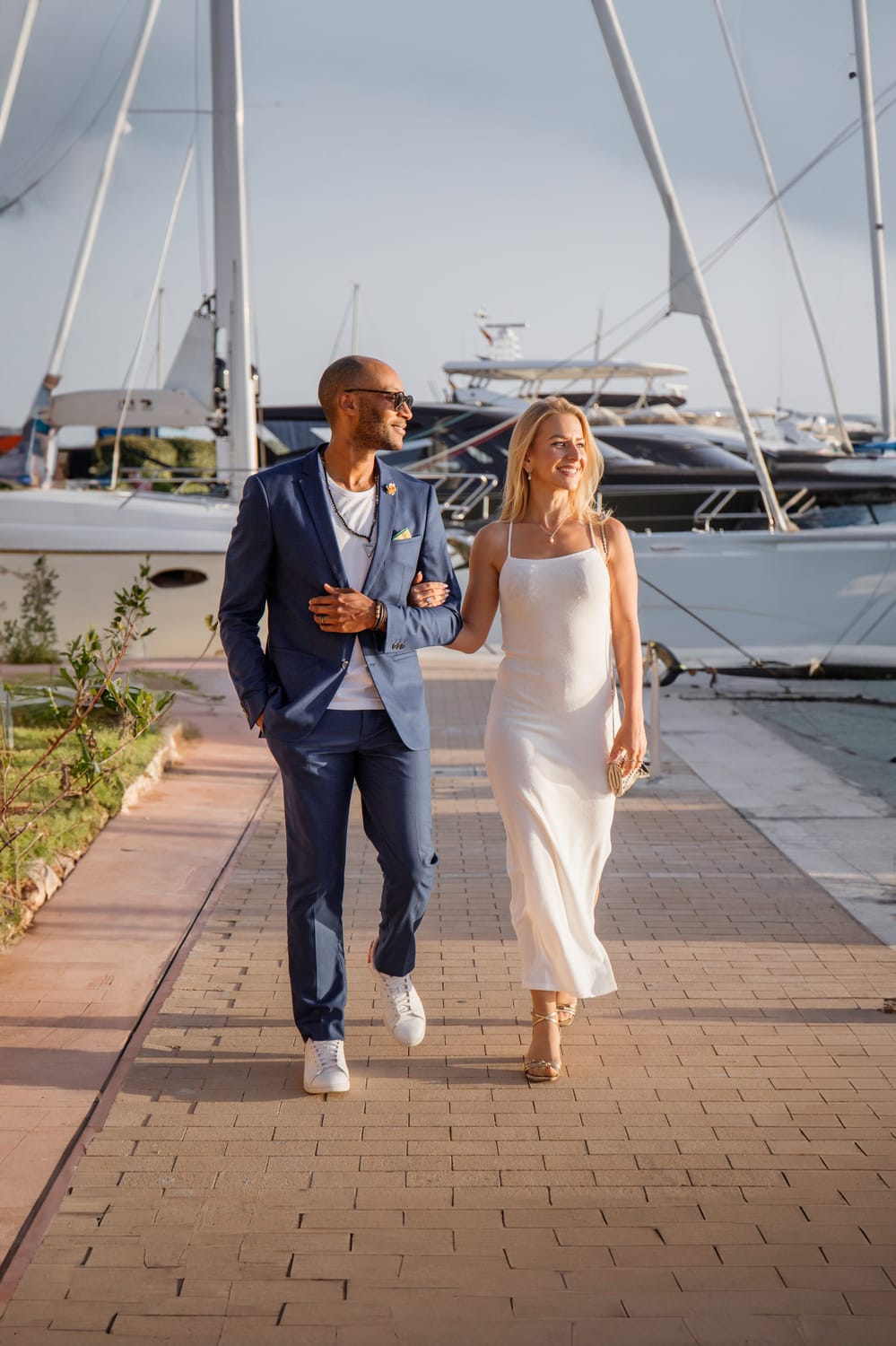 A man in a blue suit walking along a marina with a woman in a long white dress. In the background are yachts.