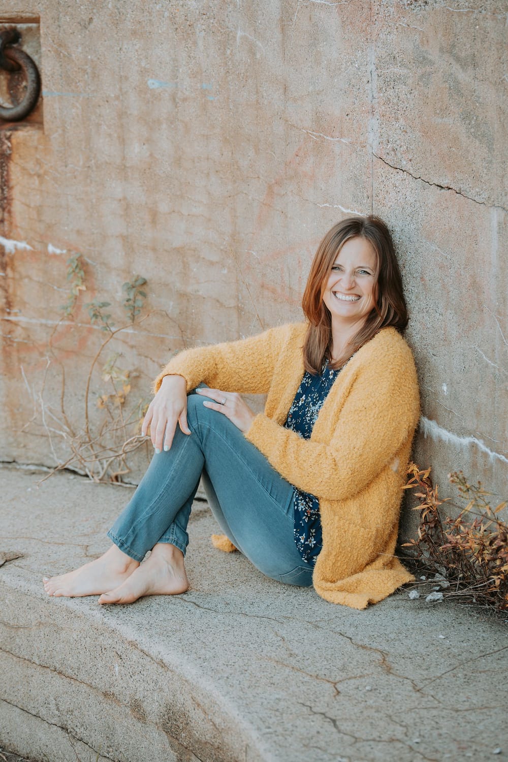 Kelly is sitting with her hand on her knee, smiling, wearing a yellow sweater.