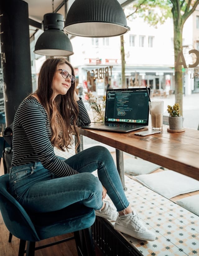Young woman working on a laptop with code visible, looking out of a café window – represents senior developers seeking a leadership path