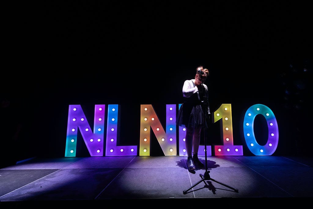Image from tenth birthday, dance commander bryony is on stage in front of an LED NLNL 10 backdrop. her hand is on her heart and she is looking humbled.