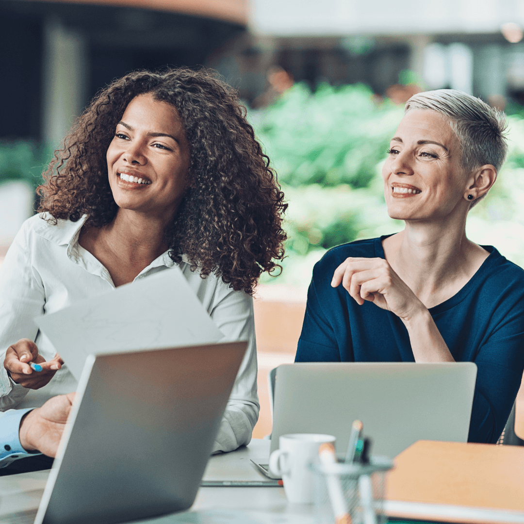 Two professional women smiling and collaborating with laptops and documents, showcasing success and teamwork in a modern, vibrant workspace. 