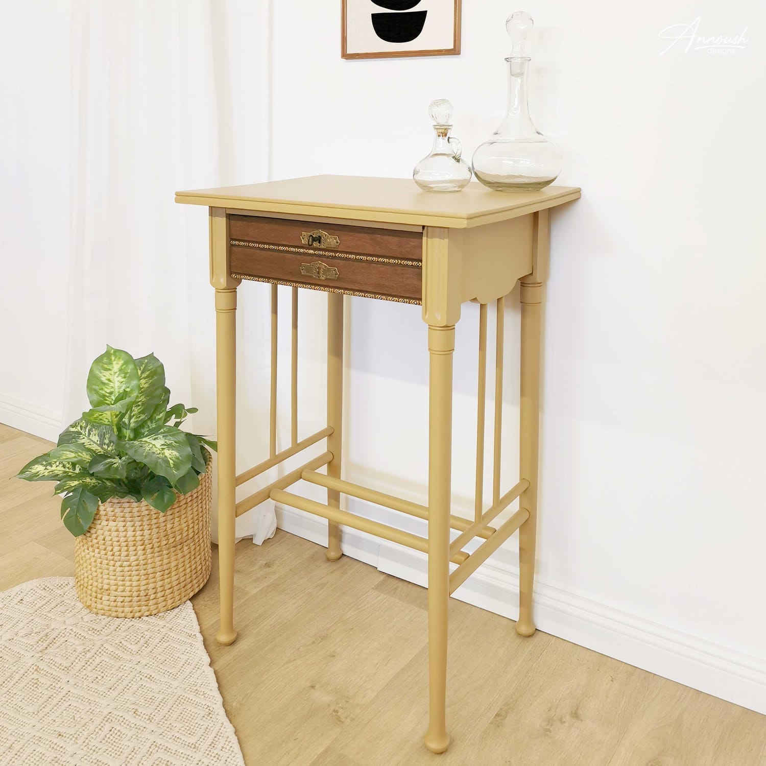 Seitenansicht des goldgelben Boho Beistelltischs im Farbton "Golden Hour", dekoriert mit zwei geschwungenen Glaskaraffen und einem geometrischen schwarz-weiß-Bild an der Wand. Davor liegt ein cremefarbener Teppich, daneben hängt ein weißer Vorhang und eine grünblättrige Pflanze im Bastkorb steht daneben.
