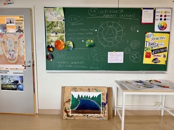 Kuvataideluokan liitutaululla ohjeita ja tehtäviä. Väriympyrän ohjeet lyhyesti 7.lk oppilaille. Taidejulisteita ja akvarellitöitä kuviluokan seinällä.