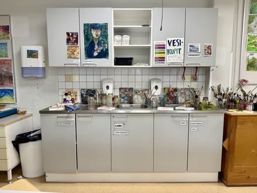 Vesipisteet kuvataideluokassa. Tiskipöydälle unohtuneet vesivärit ja lojuvat siveltimet ovat aerkea koulun kuvisluokassa. Taidejulisteita ja taidekortteja luokan seinällä ja kaappien ovissa.