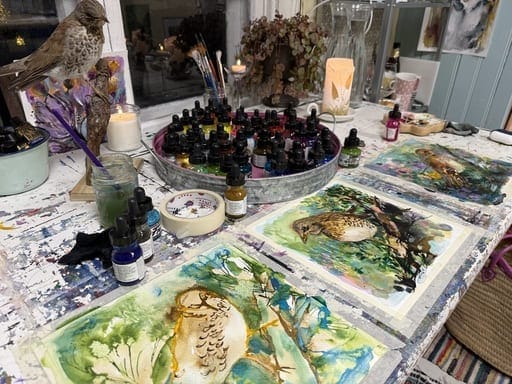 Lintuaiheisia maalauksia ateljeen pöydällä. Akryylimustepulloja ja mallina täytetty lintu. Kynttilä luo tunnelmaa pöydällä.