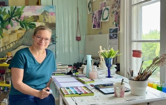 Ulla istuu altejeella työpöydön ääressä. Pöydällä Kuretake-väreja, akvarellivärit ja siveltimiä purkissa. Taustalla akryylimaalaus.