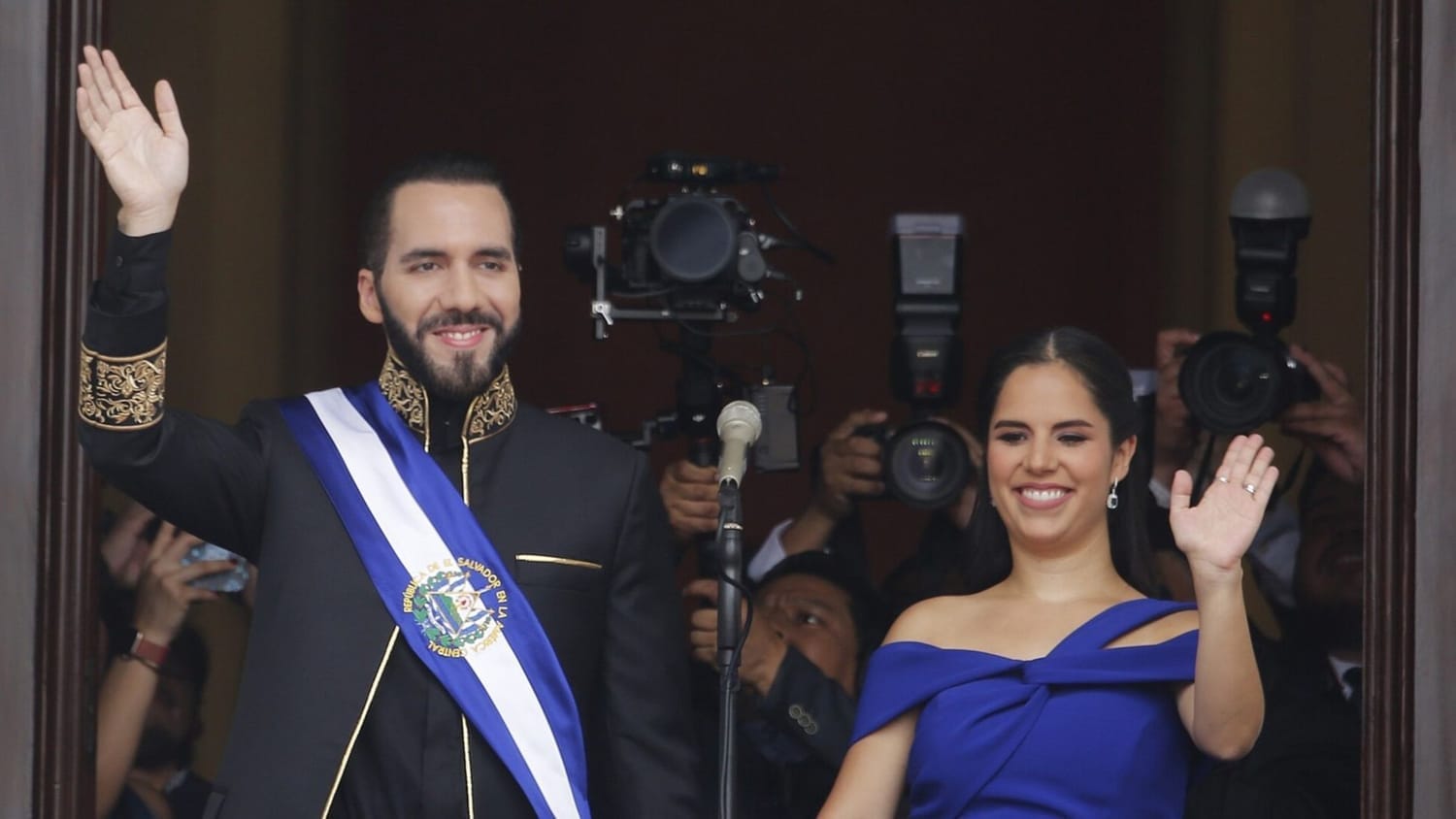 President Nayib Bukele and First Lady Gabriela Rodríguez wave to the public during Bukele’s presidential inauguration in El Salvador, with media photographers capturing the historic political event.