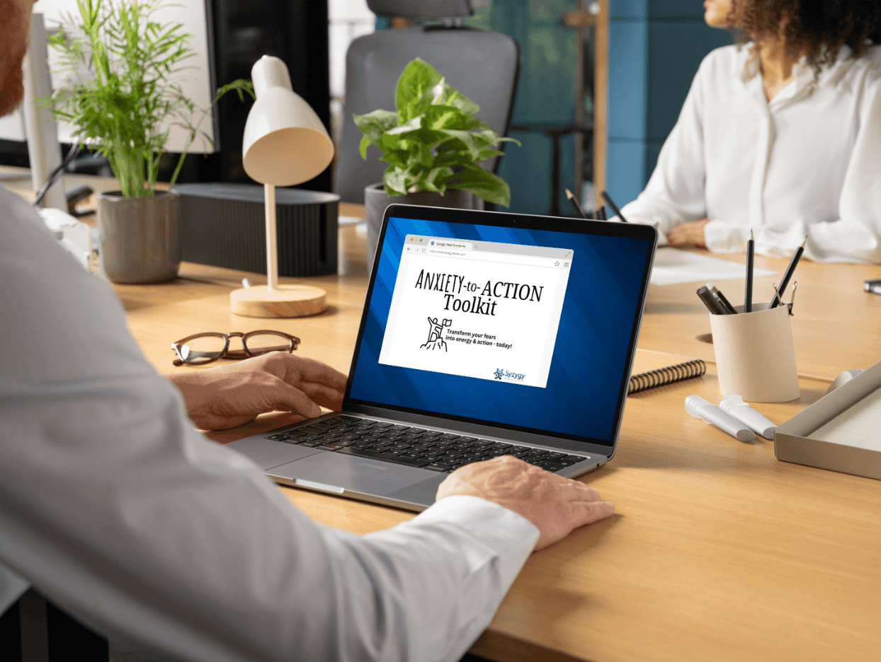 Laptop computer on a light wood desk with a notepad and pens nearby. Laptop screen showing title page of Toolkit - Anxiety-to-Action Toolkit: Transform your fears into energy and action - today!