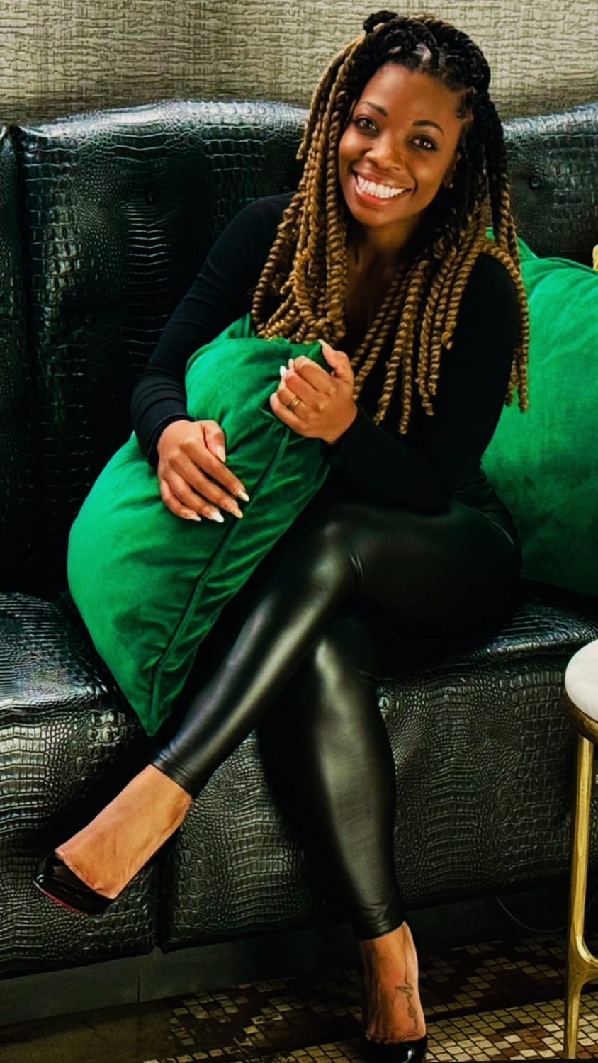 Kala C. Simmons, MSW | Confidence & Authenticity Coach, Visionary Founder & CEO of The Kollective, smiling wide. seated on a black leather couch surrounded with plush green pillows