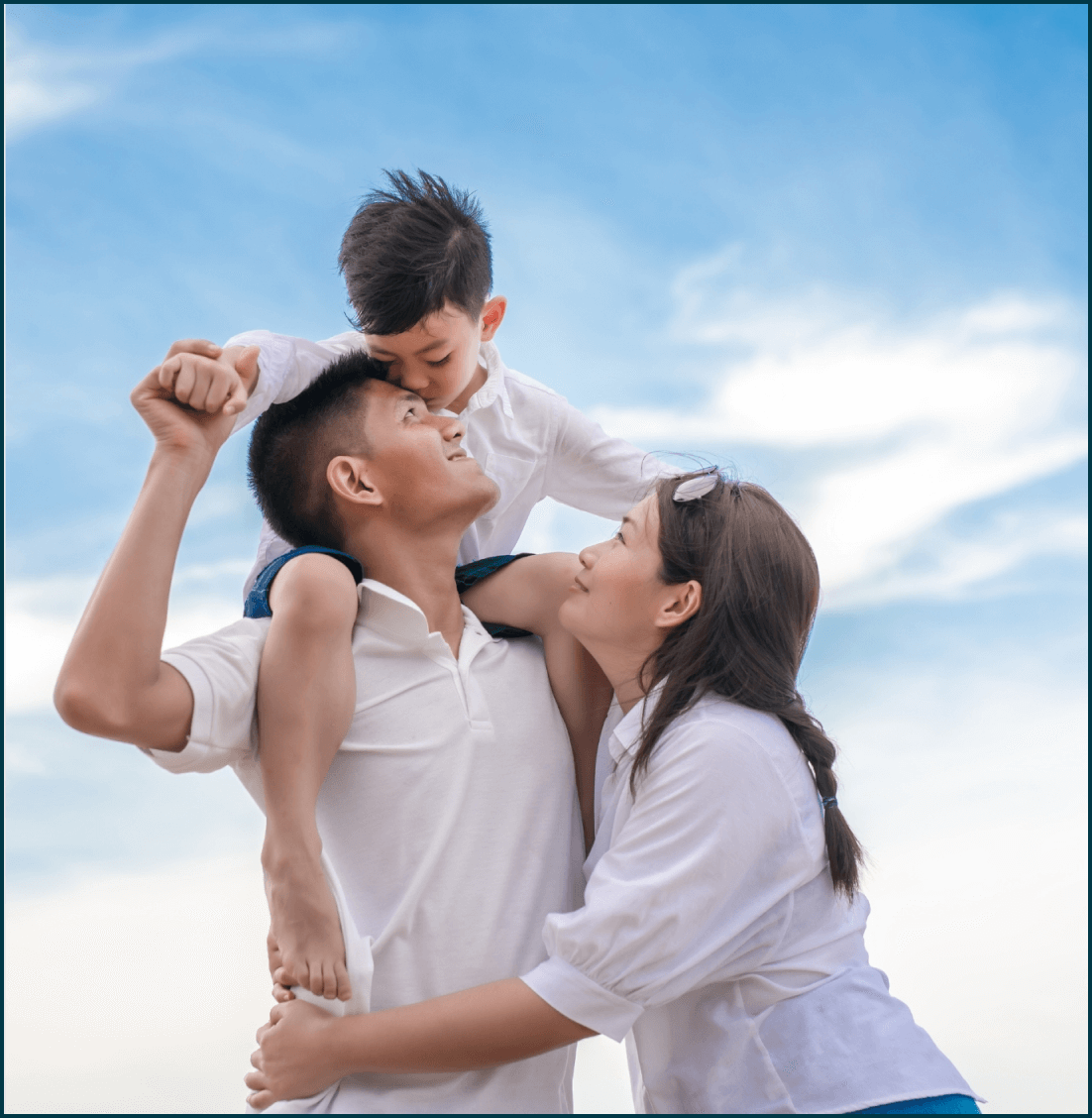 Young Filipino family smiling outdoors, child on dad’s shoulders, mom hugging. Love and security.