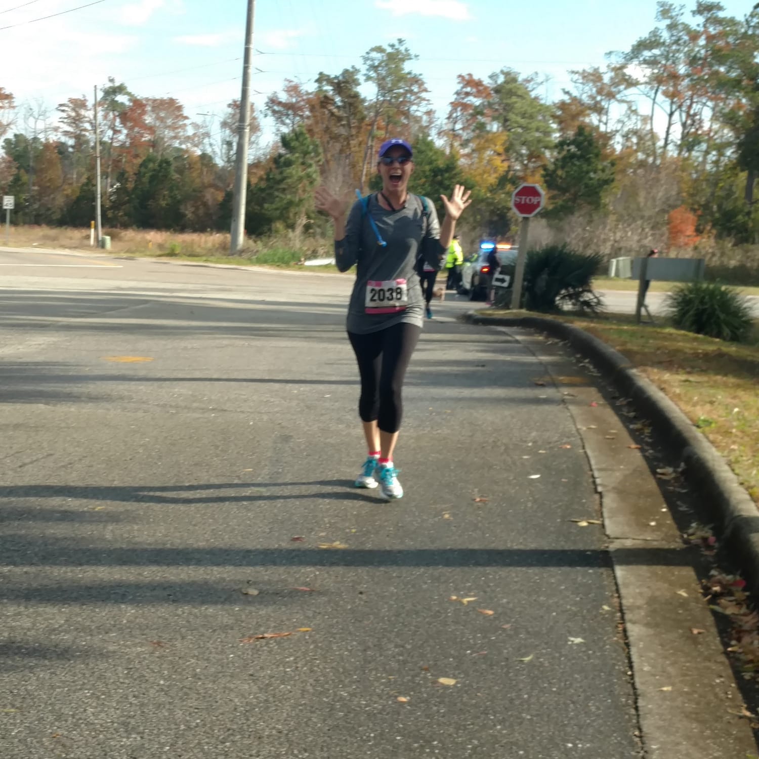 women in her 30's running, wearing leggins and a longsleeve shirt, this woman has on a race bib, a camel back and a hat. The women's hands are thrown up in joy